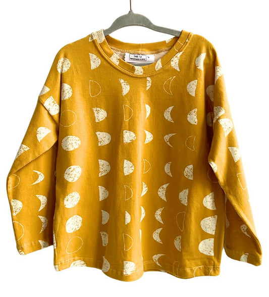 This image features a long sleeve t-shirt presented on a hanger, displaying a vibrant mustard yellow color adorned with a playful white moon and dots pattern. The t-shirt is crafted from 100% cotton, ensuring both comfort and durability.